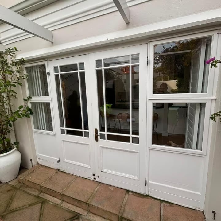 Modern white glass double doors and windows leading to a bright conservatory or sunroom with tiled flooring and potted plants.