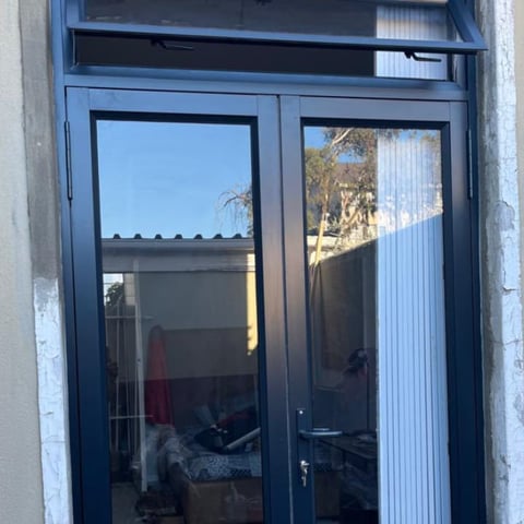 Blue-framed double glass doors with transom window, reflecting sky and surrounding structures, with white concrete walls visible on sides