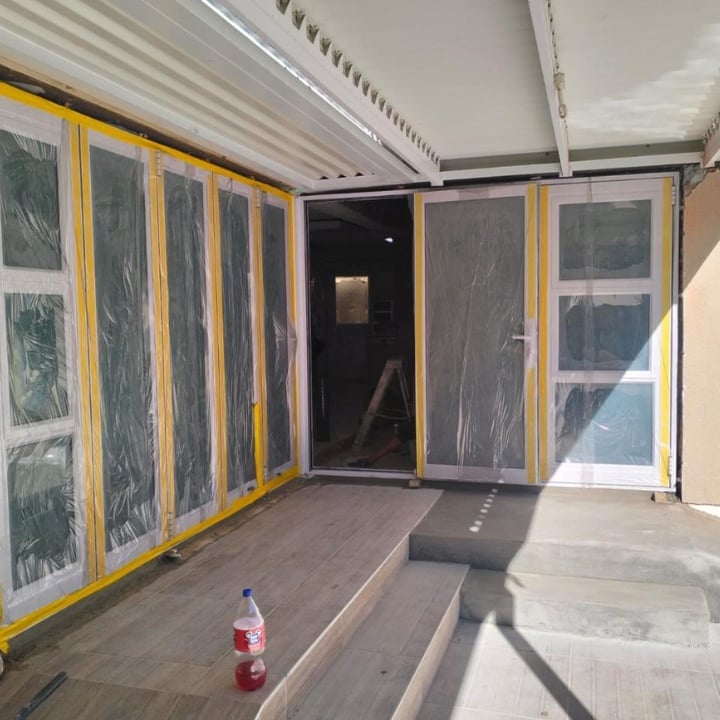 Interior of a construction site trailer with yellow-framed windows, plastic sheeting, wooden flooring, and a beverage bottle on the floor
