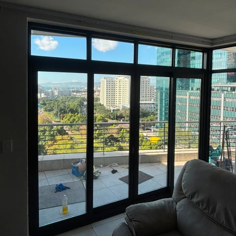 Black framed sliding glass doors and windows from an apartment showing a cityscape with buildings, trees, and sky.