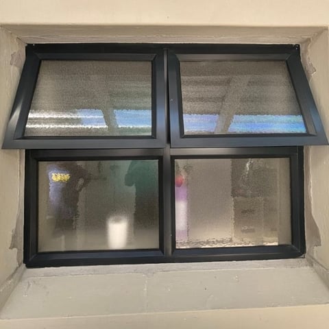 Four-pane window with black frames set in cream-colored wall, upper panes showing cloudy sky view, lower panes with condensation and reflections