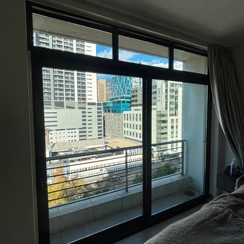 Floor-to-ceiling glass sliding doors and windows overlooking a cityscape with tall buildings, metal railing on balcony, and gray curtains