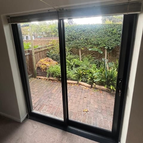 Black framed glass sliding doors opening to a brick patio garden with green hedge, wooden fence, and planted areas.