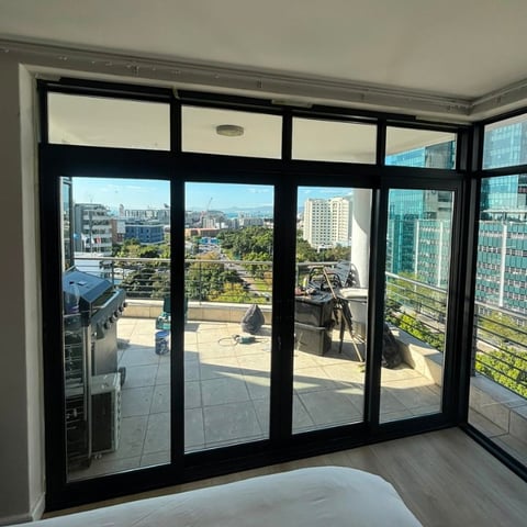 Modern apartment interior with black-framed glass doors and windows overlooking an urban skyline with tall buildings and a rooftop terrace