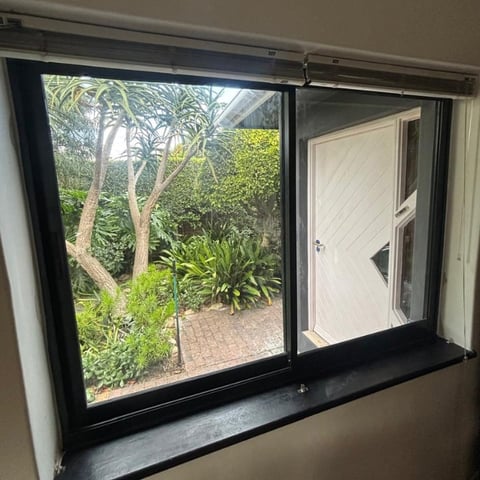Interior view through large window showing lush green garden with trees, plants, and white door outside on brick patio