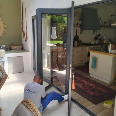 Man installing or maintaining a black-framed glass bifold door in a kitchen with light green walls and wooden cabinets