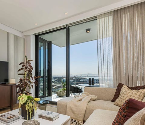 Modern high-rise apartment living room with floor-to-ceiling windows overlooking cityscape, beige sofa, plants, and sheer curtains