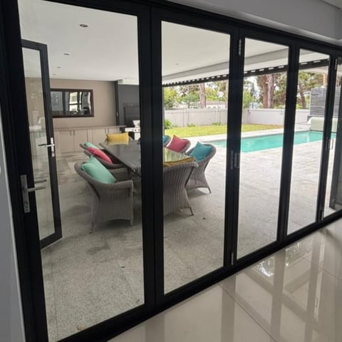 Black frame sliding glass doors opening to a patio with wicker chairs, cushions, and a swimming pool beyond
