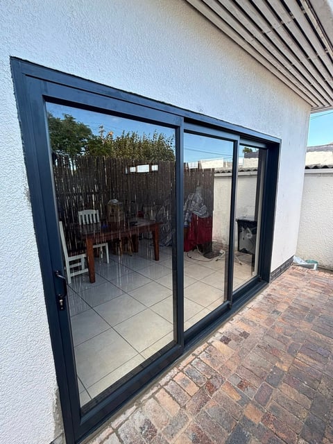 Modern black framed sliding glass doors opening onto a patio with red chairs and potted plants against a white building wall.