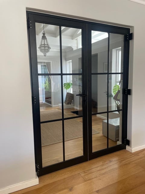 Black framed double glass doors with grid panes opening into a bright interior room with wooden flooring