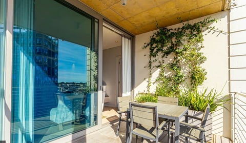 Modern balcony with table and chairs, climbing vines, and floor-to-ceiling windows overlooking a city waterfront