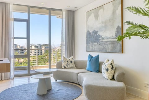 Modern living room with curved gray sofa, blue accent pillows, round coffee table, floor-to-ceiling windows overlooking cityscape, and potted fern plant