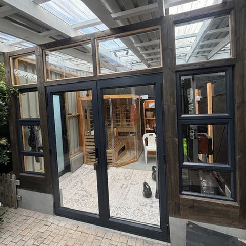 Modern black-framed glass conservatory with skylight ceiling and glazed doors leading to an interior wellness spa featuring a wooden sauna